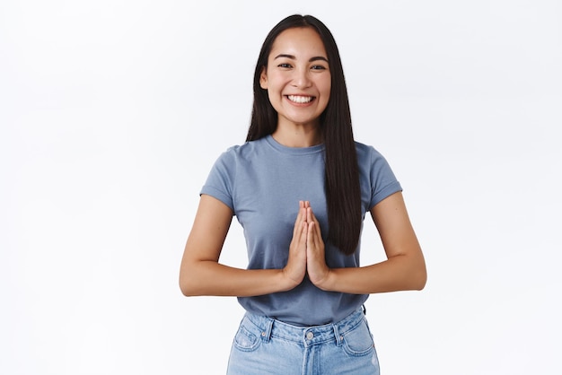 Linda y agradable mujer morena asiática con jeans de camiseta sonriendo ampliamente toman las manos para rezar sobre el pecho agradeciendo la ayuda que parece agradecida expresar gratitud o rezar cerca del fondo blanco del santuario