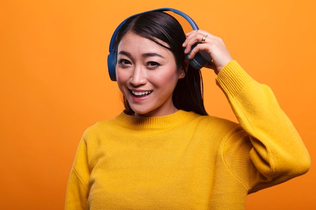 Una linda y alegre persona asiática positiva deja de escuchar música para hablar con un compañero mientras está en un fondo naranja. Hermosa mujer sonriente de corazón quitándose los auriculares cuando conoce a su amiga.