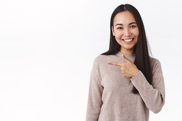 Linda amiga asiática da consejos sobre dónde comprar para las vacaciones, recomienda un enlace o una aplicación, apunta a la izquierda y sonríe alegremente, de pie amistosamente sobre una pared blanca, tiene una discusión informal