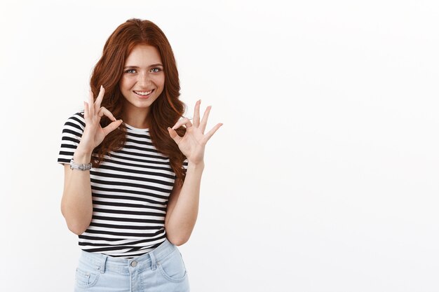 Linda y amistosa adolescente pelirroja en camiseta a rayas, da su aprobación, totalmente como tu elección, sonriendo satisfecho, muestra un gesto de permiso aceptable, juzgando una idea excelente, pared blanca