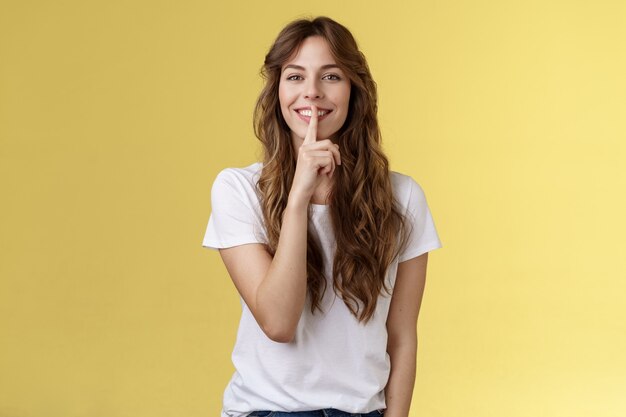 Linda astucia encantadora chica europea peinado rizado ocultando el secreto de la belleza sonriendo sensualmente mostrar silencio silencio gesto dedo índice presionó los labios sonriendo con alegría de pie fondo amarillo.