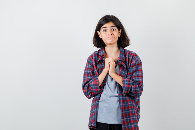 Linda chica adolescente juntando las manos en gesto de oración en camisa a cuadros y mirando confundido, vista frontal.