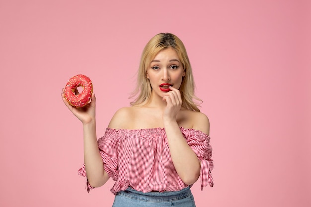 Foto gratuita linda chica adorable chica rubia con lápiz labial rojo en blusa rosa mordiendo el dedo sosteniendo una rosquilla