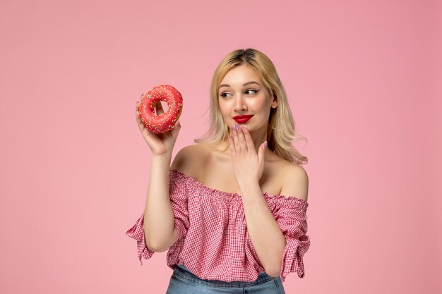 Linda chica adorable chica rubia con lápiz labial rojo en blusa rosa que se ve encantadora en una rosquilla de bayas