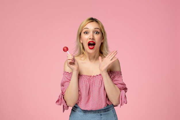 Linda chica adorable chica rubia con lápiz labial rojo en blusa rosa tan emocionada por una piruleta