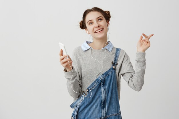 Linda chica adulta con el pelo en bollos dobles sonriendo escuchando música desde su teléfono móvil gesticulando con placer. Persona femenina contenta bailando escuchando melodías con auriculares. Concepto de pasatiempo