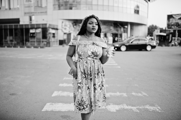 Foto gratuita linda chica afroamericana de pequeña altura con rastas vestidas con un vestido amarillo de color caminando en el cruce de peatones contra el centro comercial con una taza de café a mano en blanco y negro