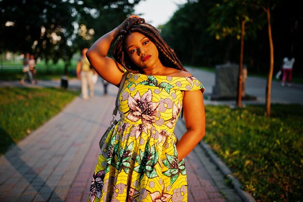 Linda chica afroamericana de pequeña altura con rastas en un vestido amarillo de color posado al atardecer