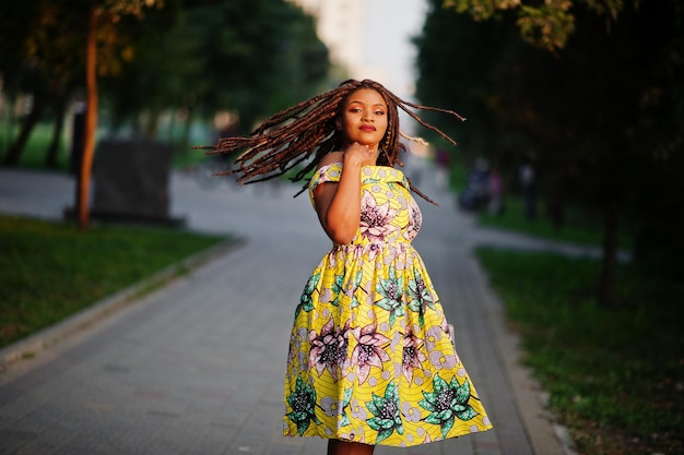 Linda chica afroamericana de pequeña altura con rastas en un vestido amarillo de color posado al atardecer