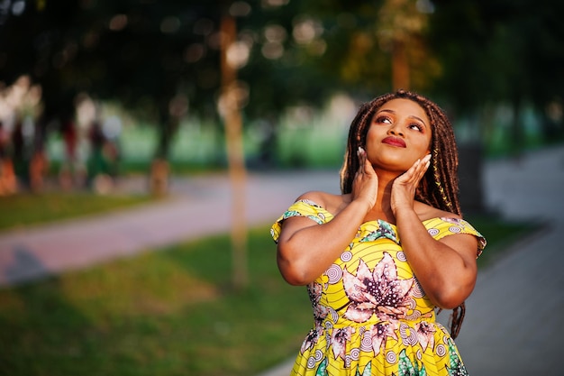 Linda chica afroamericana de pequeña altura con rastas en un vestido amarillo de color posado al atardecer