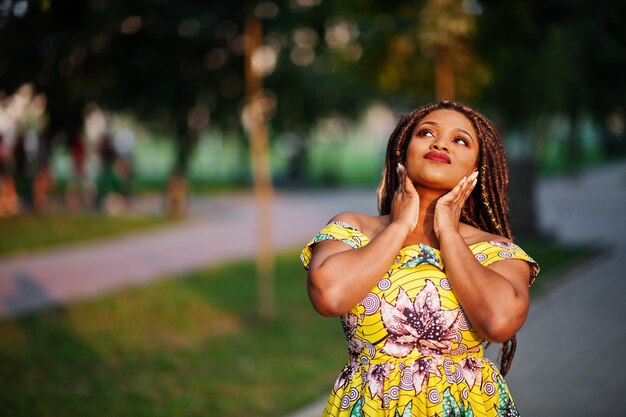 Linda chica afroamericana de pequeña altura con rastas en un vestido amarillo de color posado al atardecer