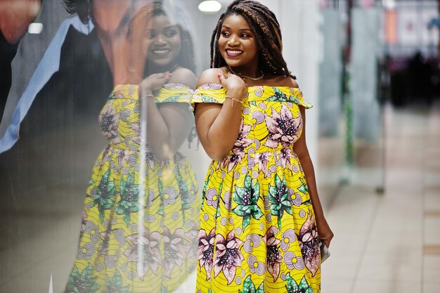 Linda chica afroamericana de pequeña altura con rastas en un vestido amarillo de color posado en el escaparate de la tienda en el centro comercial