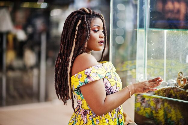 Linda chica afroamericana de pequeña altura con rastas en un vestido amarillo de color que mira el acuario en el centro comercial