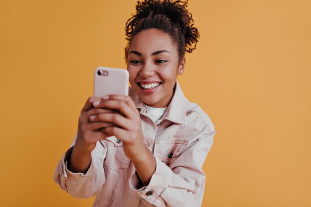 Linda chica afroamericana tomando fotos con un teléfono inteligente Vista frontal de una impresionante dama negra usando un dispositivo digital sobre fondo naranja