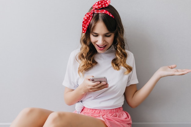 Linda chica alegre en camiseta blanca mirando la pantalla del teléfono. Señora sonriente extática con peinado ondulado posando en casa con smartphone.