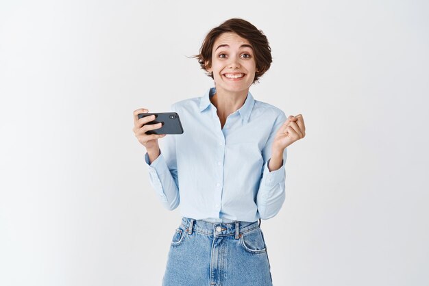 Linda chica alegre ganando en el teléfono móvil haciendo dinero en la aplicación de teléfono inteligente y sonriendo de pie feliz sobre fondo blanco