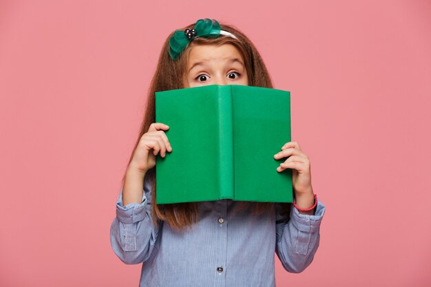 Linda chica con aro de pelo divirtiéndose mientras lee un libro interesante con los ojos bien abiertos