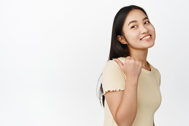 Linda chica asiática apuntando a la izquierda y sonriendo mostrando el banner de venta de la tienda detrás de ella de pie en camiseta sobre fondo blanco.