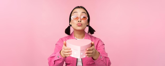 Linda chica asiática dándote un regalo en una caja con forma de corazón besándose y sonriendo concepto de fiesta y celebración de pie sobre fondo rosa