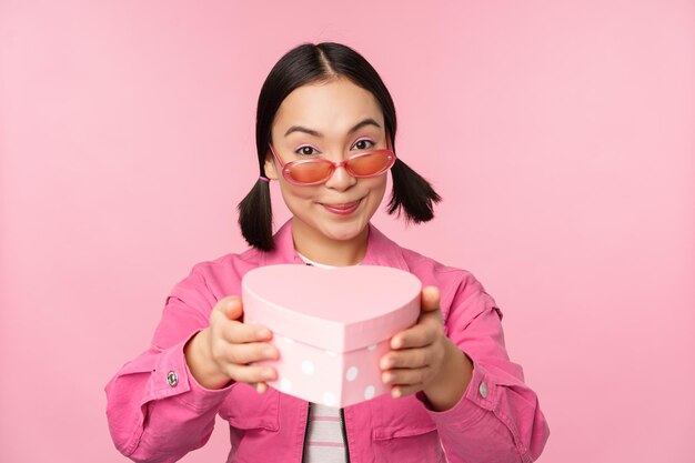 Linda chica asiática dándote un regalo en una caja con forma de corazón besándose y sonriendo concepto de fiesta y celebración de pie sobre fondo rosa