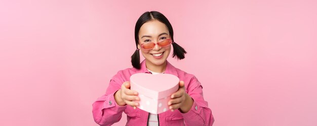 Linda chica asiática dándote un regalo en una caja con forma de corazón besándose y sonriendo concepto de fiesta y celebración de pie sobre fondo rosa