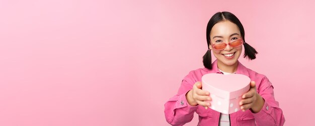 Linda chica asiática dándote un regalo en una caja con forma de corazón besándose y sonriendo concepto de fiesta y celebración de pie sobre fondo rosa