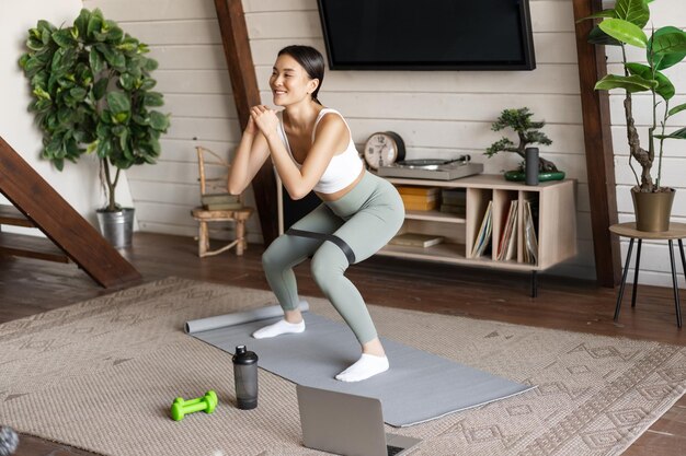 Linda chica asiática fitness en casa haciendo sentadillas de entrenamiento con estiramiento de cuerda elástica en las piernas de pie o ...