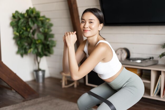 Linda chica asiática fitness en casa haciendo sentadillas de entrenamiento con estiramiento cuerda elástica en sus piernas ...