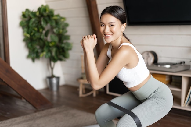 Linda chica asiática fitness en casa haciendo sentadillas de entrenamiento con estiramiento cuerda elástica en sus piernas ...