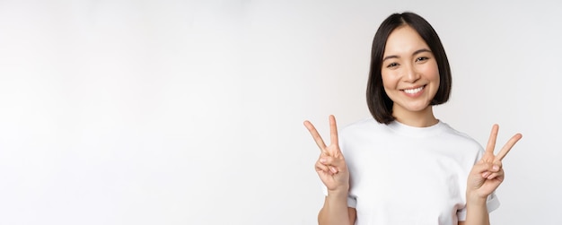 Linda chica asiática mostrando signo de paz sonriendo y mirando feliz a la cámara con estudio de camiseta blanca