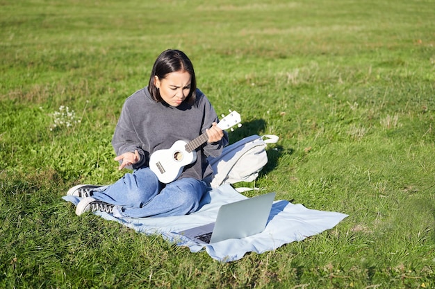 Linda chica asiática músico estudiando mira tutorial en su computadora portátil tutor en línea muestra cómo tocar uku