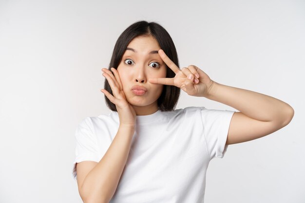 Linda chica asiática posando con kawaii vsign gesto de paz cerca de la cara de pie en camiseta sobre fondo blanco Copiar espacio