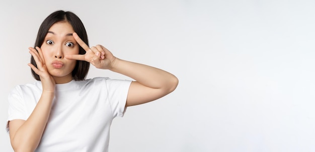 Linda chica asiática posando con kawaii vsign gesto de paz cerca de la cara de pie en camiseta sobre fondo blanco Copiar espacio