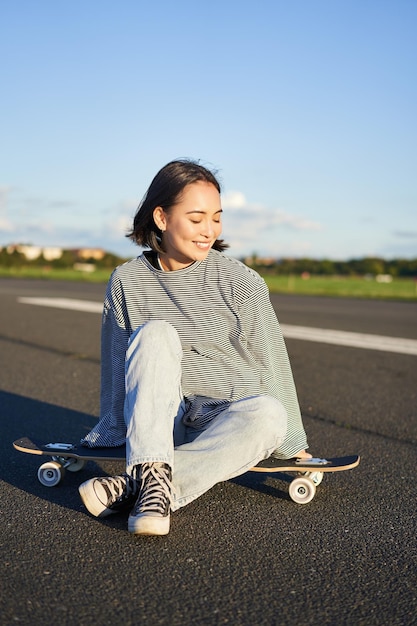 Una linda chica asiática sonriente se sienta sola en un monopatín de longboard en un espacio de copia de carretera vacío