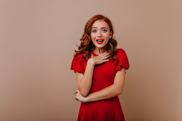 Linda chica asombrada con pelo largo oscuro de pie en la pared clara. Mujer europea refinada en la presentación del vestido rojo.