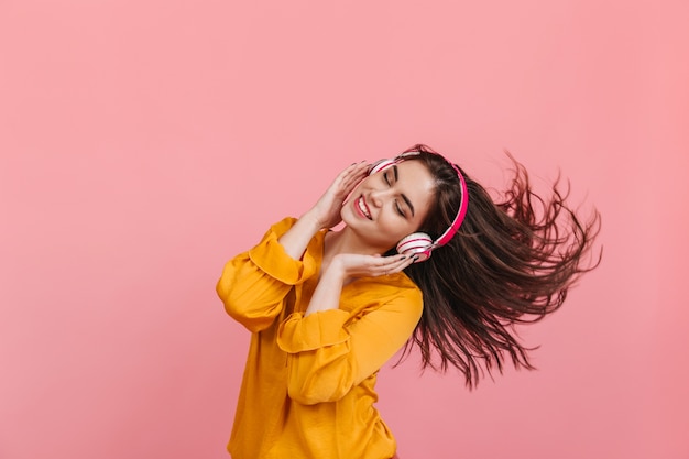 Linda chica en auriculares blancos y rosas bailando activamente en la pared rosa, moviendo el pelo largo.