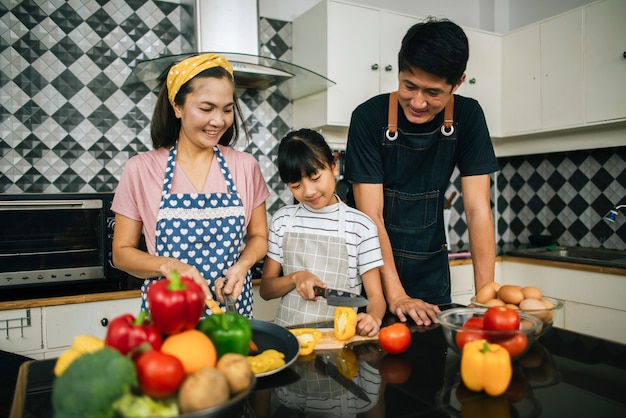 Linda chica ayuda a sus padres a cortar vegetales y sonreír mientras cocinan juntos en la cocina