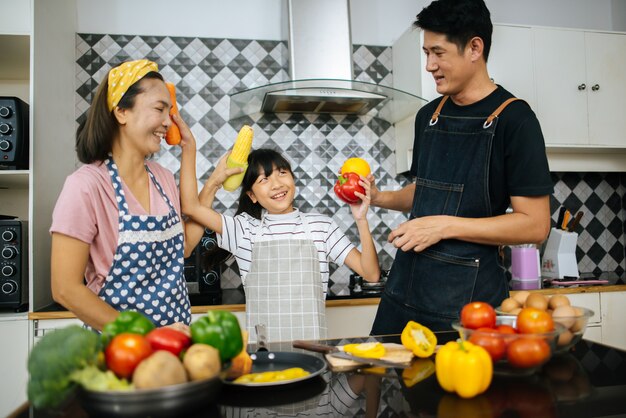 Linda chica ayuda a sus padres a cortar vegetales y sonreír mientras cocinan juntos en la cocina