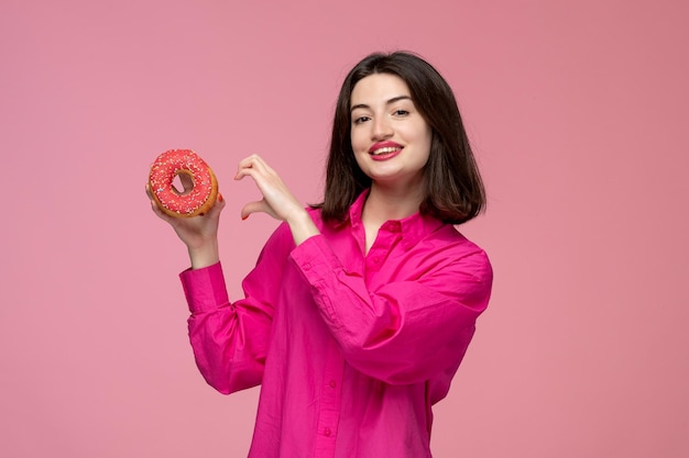 Linda chica bastante adorable chica en camisa rosa con lápiz labial rojo mostrando signo de corazón a donut