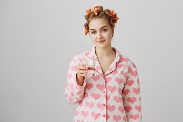 Linda chica bebiendo agua antes de dormir, usa pijamas y rizadores de pelo