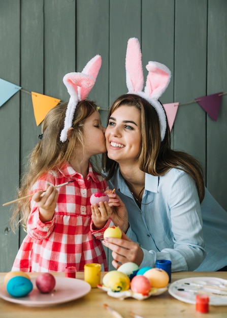 Linda chica besando a la madre en la mejilla mientras pinta huevos para Pascua