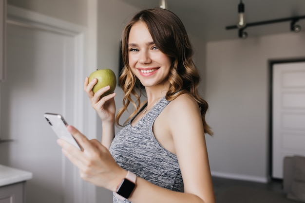 Linda chica bien formada posando con manzana verde en casa. Foto interior de mujer rizada dichosa sosteniendo smartphone con sonrisa.