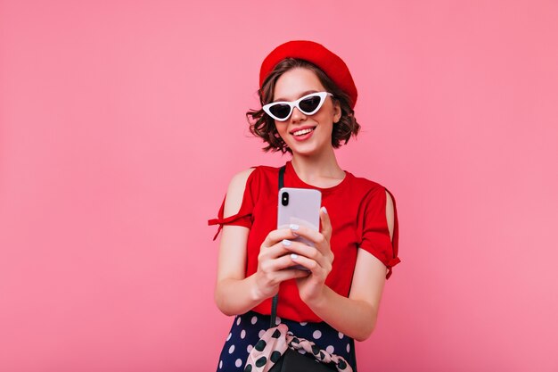 Linda chica blanca de boina roja tomando una foto de sí misma con una sonrisa. Foto interior de magnífica mujer de pelo corto con gafas de sol haciendo selfie.