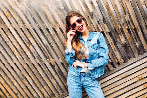 Linda chica blanca en traje de estilo retro riendo y bromeando. Hermosa joven sonriente con cabello largo aislado sobre un fondo de valla de madera.