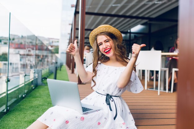 Linda chica con cabello largo está sentada en el piso de la terraza. Lleva un vestido blanco con hombros desnudos, lápiz labial rojo y sombrero. Ella tiene una computadora portátil de rodillas y sonríe a la cámara.