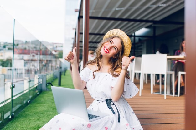 Linda chica con cabello largo con sombrero está sentada en el piso de la terraza. Lleva un vestido blanco con hombros desnudos, lápiz labial rojo. Tiene una computadora portátil sobre las rodillas y parece afortunada.