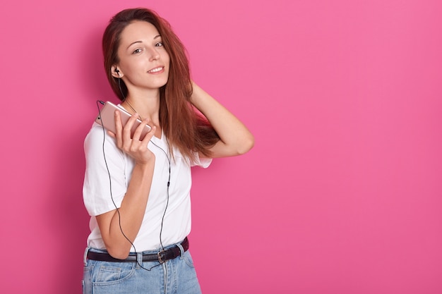 Linda chica de cabello oscuro escuchando música a través de auriculares en el teléfono inteligente, divirtiéndose, vistiendo una camiseta blanca y jeans, mantiene la mano detrás de la cabeza