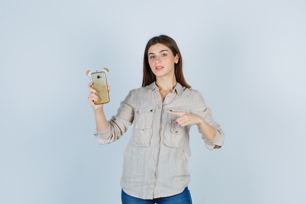 Linda chica en camisa apuntando al teléfono móvil y mirando confiada