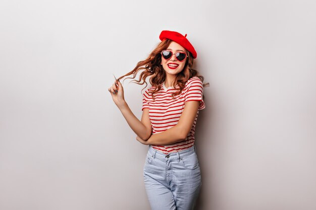 Linda chica caucásica en jeans y boina de pie en la pared blanca. Foto interior de risa elegante joven con cabello pelirrojo.