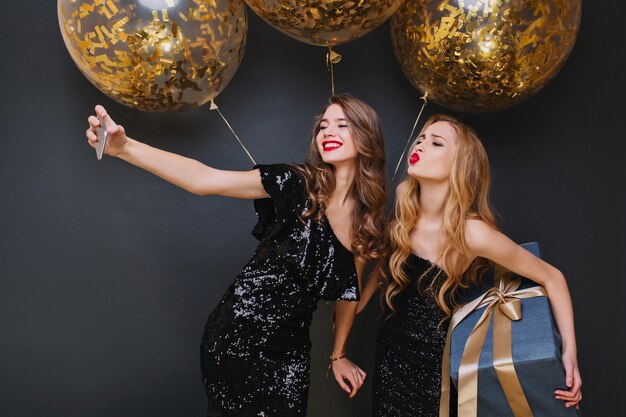 Linda chica caucásica con pelo largo y rizado posando con expresión de cara de besos, sosteniendo un gran regalo. Mujer joven relajada haciendo selfie con un amigo durante la celebración de la Navidad.